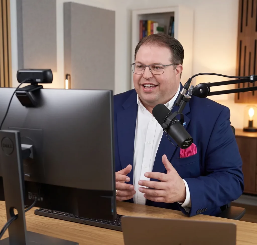 Ein Trainer sitzt lachend am Schreibtisch vor einem großen Monitor und Mikrofon für das Online-Coaching.